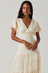 A woman is wearing a light-colored, embroidered dress with a V-neckline. The dress features short, ruffled sleeves and a fitted waist, with intricate lace and floral embroidery details throughout. The skirt is tiered with a gathered, ruffled hem.